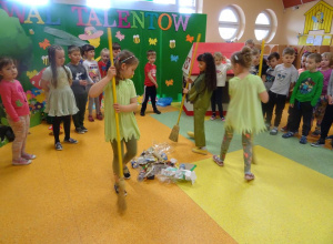 Trzy dziewczynki trzymają miotły w ręku i zamiatają rozrzucone odpady na podłodze. Wokół nich stoją dzieci, które śpiewają piosenkę.